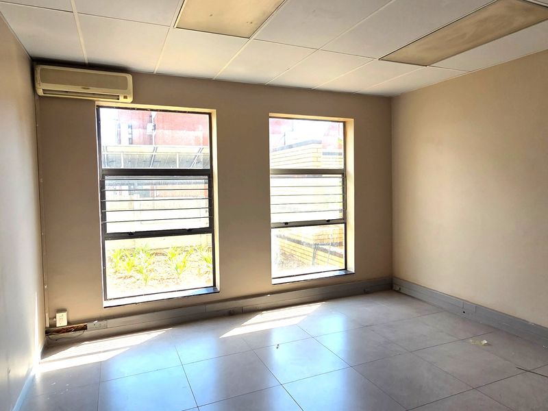An empty office space featuring tiled flooring, a suspended ceiling, and a wall-mounted air conditioning unit with natural light from two windows.
