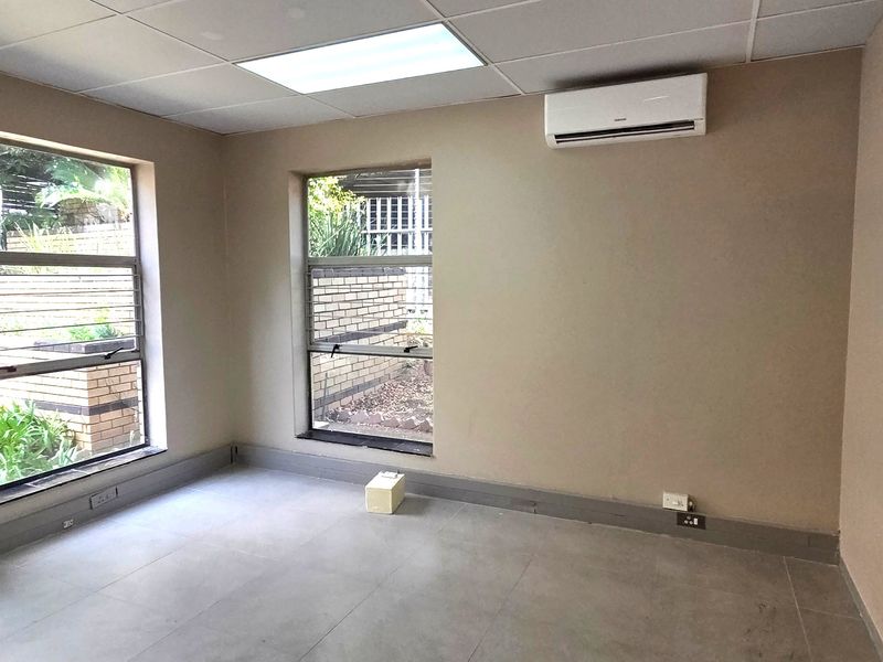 A clean, empty office space featuring tiled flooring, a suspended ceiling with recessed lighting, and a wall-mounted split-system air conditioner.