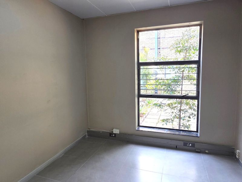 A vacant office space featuring neutral tiled flooring, electrical power skirting, and a large window with security bars, offering a clean, functional workspace.