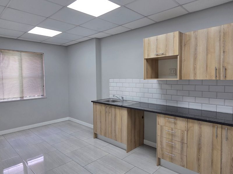 A clean, modern office kitchenette featuring light wood-grain cabinetry, dark countertops, subway tile backsplash, and bright recessed ceiling lighting.