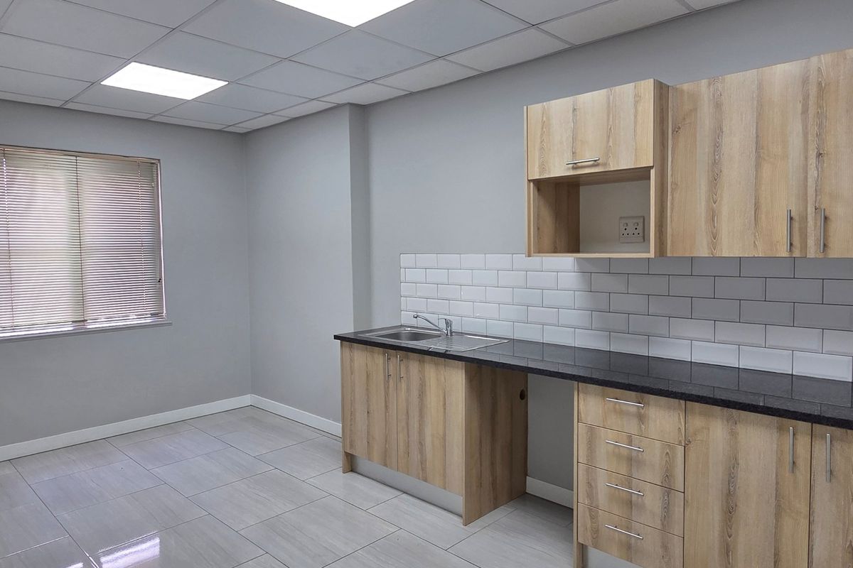 A clean, modern office kitchenette featuring light wood-grain cabinetry, dark countertops, subway tile backsplash, and bright recessed ceiling lighting.