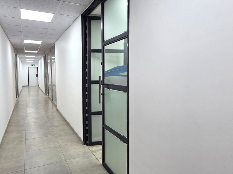 A clean, modern office hallway featuring tiled flooring, suspended ceiling with recessed lighting, and contemporary glass partition doors.
