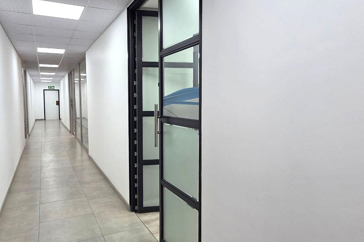 A clean, modern office hallway featuring tiled flooring, suspended ceiling with recessed lighting, and contemporary glass partition doors.