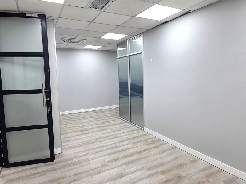 Modern, empty office space featuring neutral grey walls, wood-look laminate flooring, suspended ceiling with LED lighting, and contemporary glass partitioning.