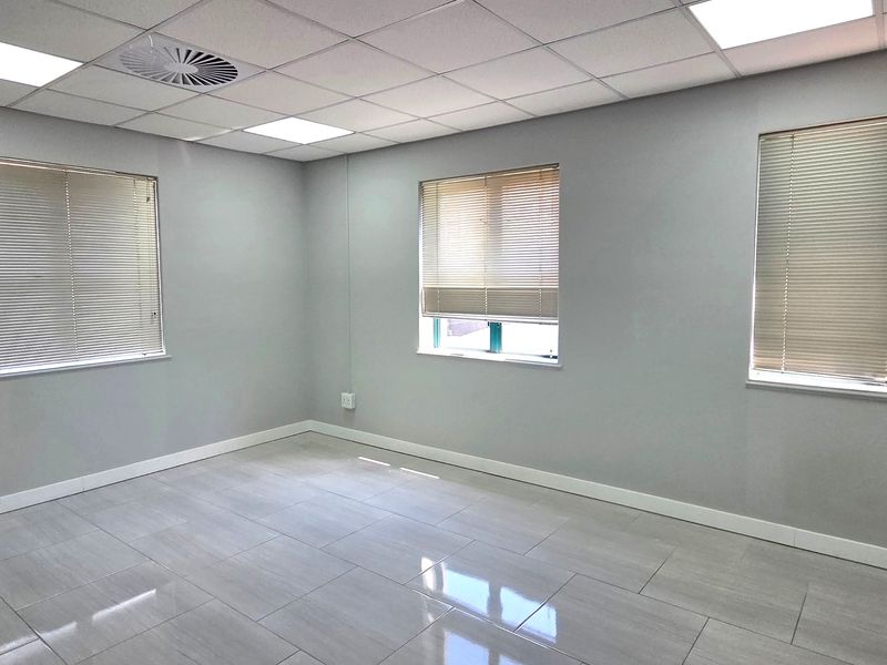 A clean, vacant office space featuring modern tiled flooring, a suspended ceiling with recessed lighting, and multiple windows fitted with venetian blinds.