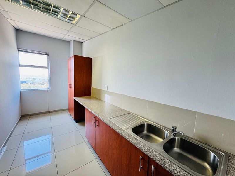 A clean, functional office kitchenette featuring built-in wooden cabinetry, a double stainless steel sink, tiled flooring, and ample counter space.