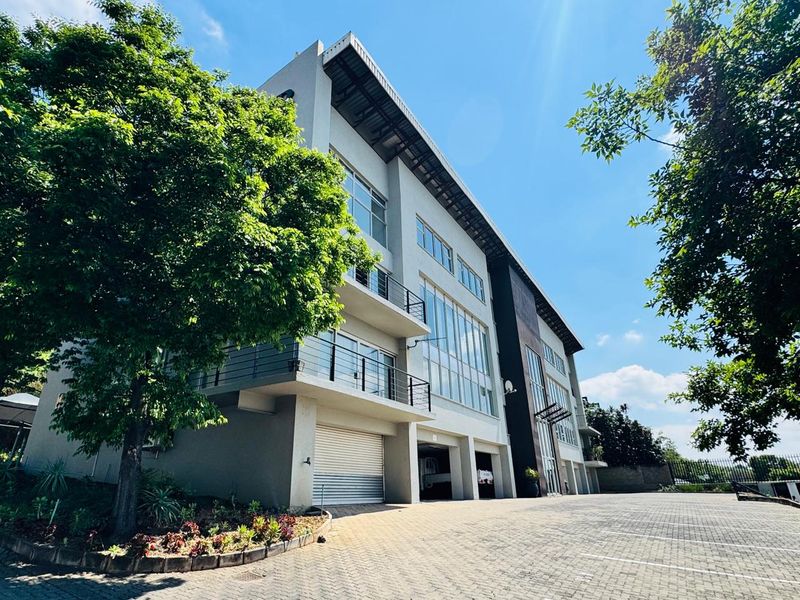 A modern multi-story office building featuring a paved driveway, large glass windows, and private balconies with metal railings.