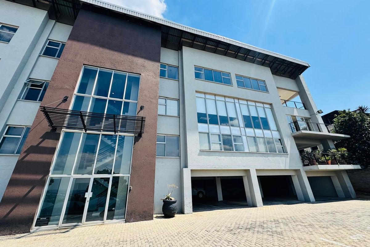Modern multi-story office building featuring a prominent glass facade, covered ground-level parking, and balconies, set against a clear blue sky.