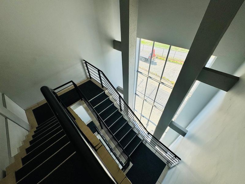 A bright, modern stairwell featuring a metal handrail, full-height windows, and exposed concrete structural columns, providing a clean and professional office aesthetic.