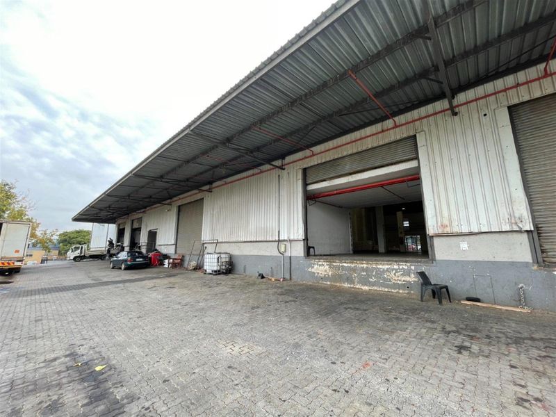 Industrial warehouse exterior featuring a large loading dock area with multiple roller shutter doors, corrugated metal cladding, and a paved yard.