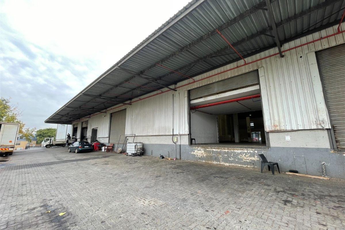Industrial warehouse exterior featuring a large loading dock area with multiple roller shutter doors, corrugated metal cladding, and a paved yard.