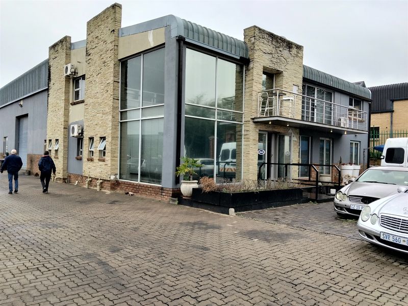 Front exterior view of a multi-level industrial building featuring stone cladding, large glass windows, a balcony, and a paved parking area.