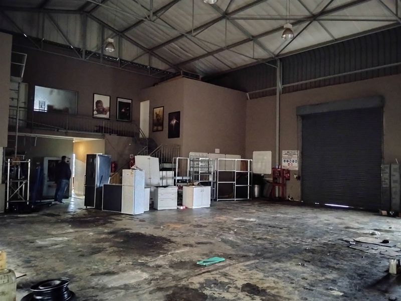 Spacious industrial warehouse featuring a high-clearance ceiling with exposed steel trusses, a large roller shutter door, and a mezzanine level with staircase access.