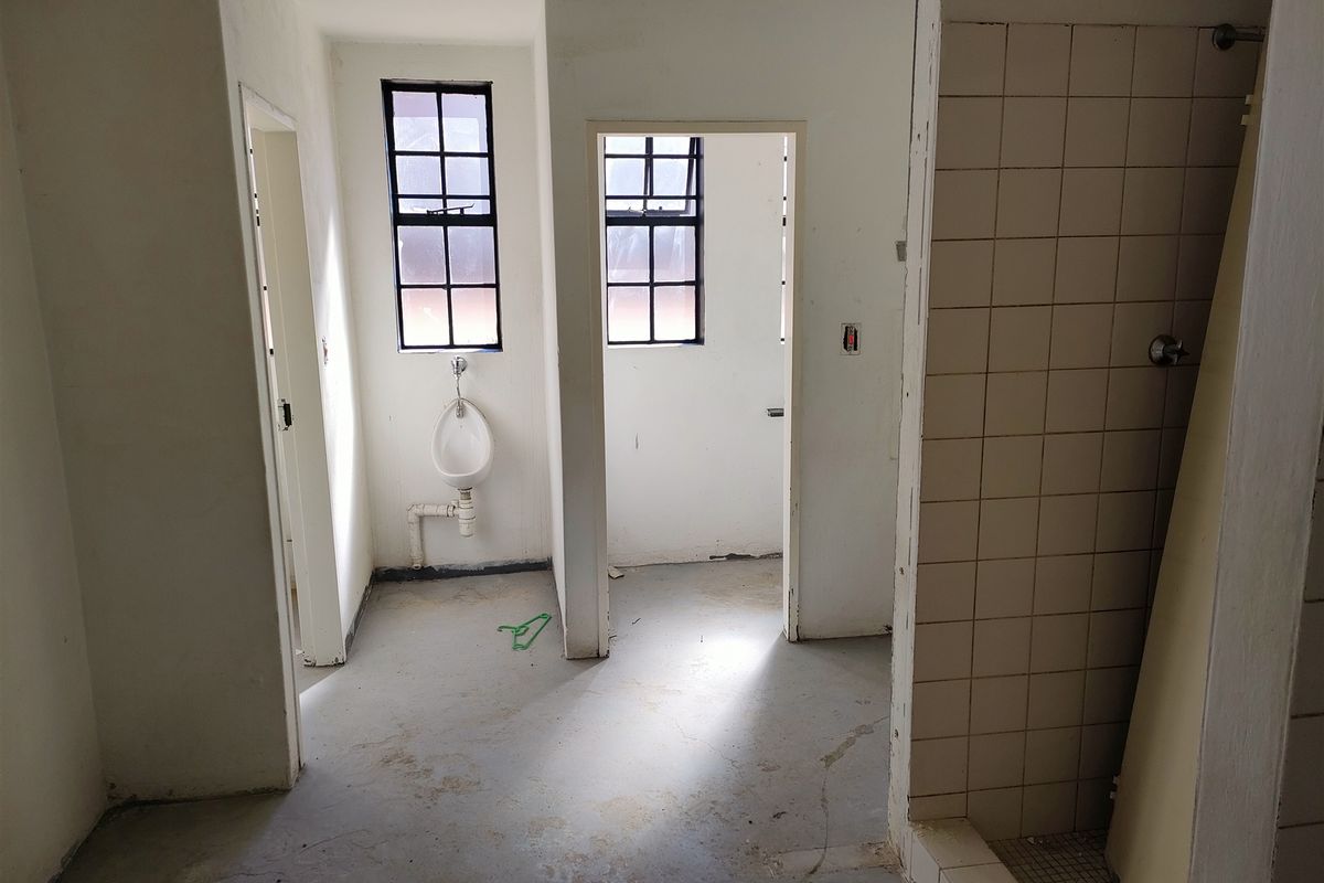 An industrial-style bathroom space featuring a wall-mounted urinal, tiled shower area, and steel-framed windows, currently in need of renovation and finishing.