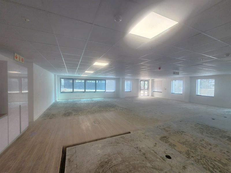 A spacious, open-plan office area featuring a suspended ceiling, recessed lighting, and large windows, currently undergoing flooring renovations.