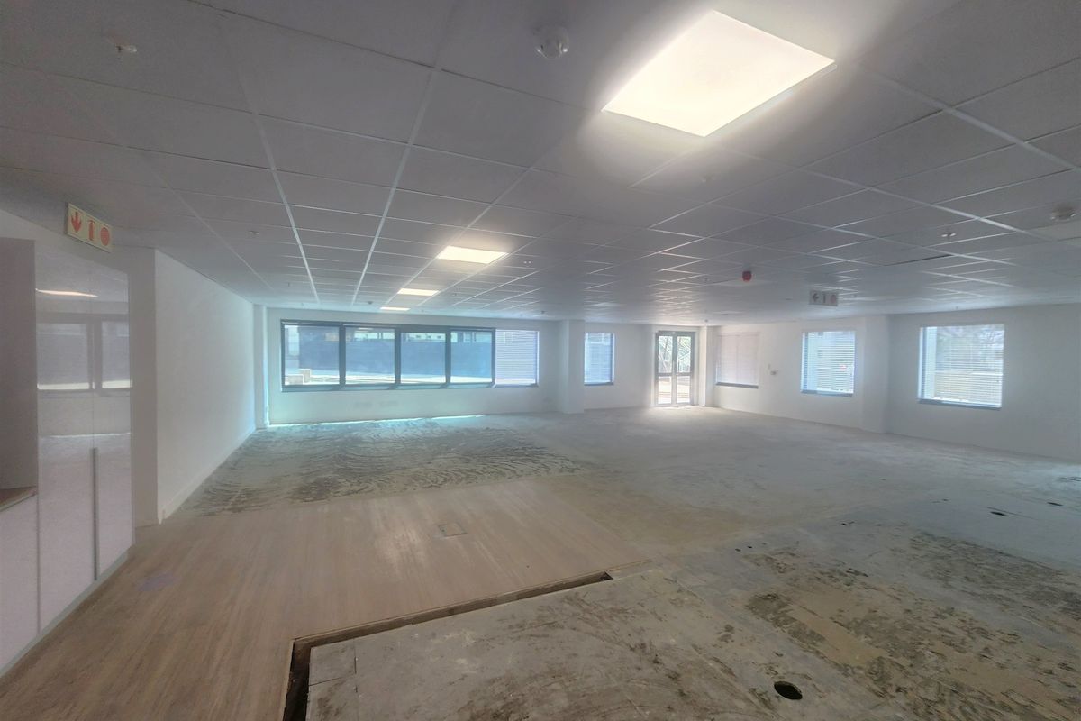 A spacious, open-plan office area featuring a suspended ceiling, recessed lighting, and large windows, currently undergoing flooring renovations.