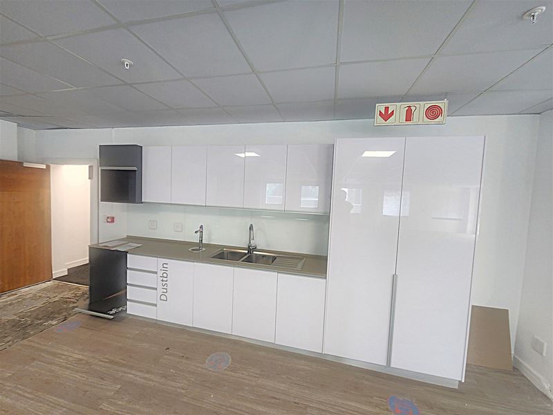 A clean, modern office kitchenette featuring white high-gloss cabinetry, a stainless steel sink, and durable laminate flooring in a professional workspace setting.