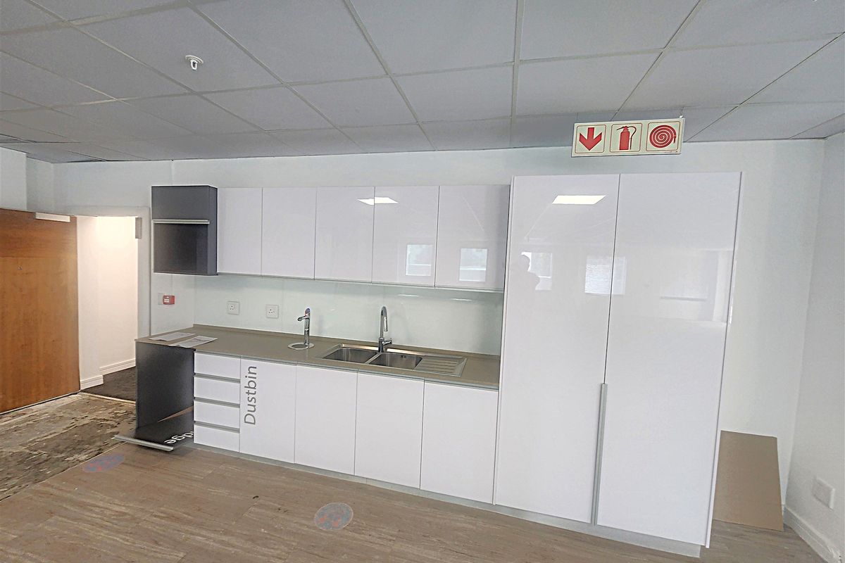 A clean, modern office kitchenette featuring white high-gloss cabinetry, a stainless steel sink, and durable laminate flooring in a professional workspace setting.