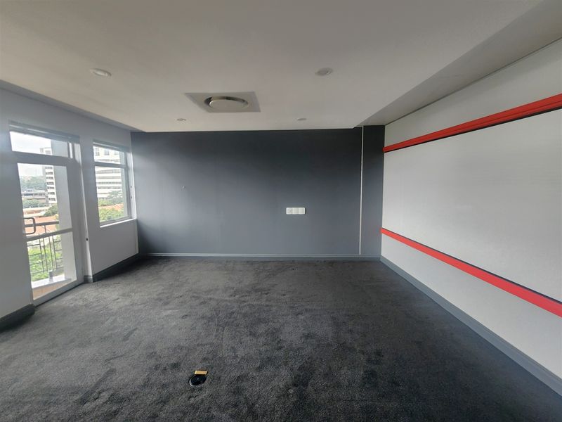 A spacious, empty office featuring dark carpeting, neutral walls with red accent trim, recessed lighting, and large windows providing natural light and city views.