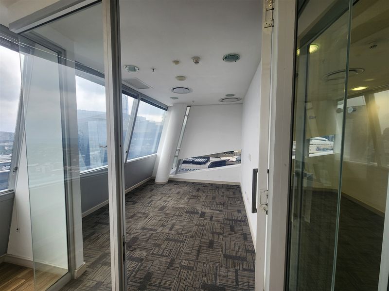 A bright, empty office space featuring floor-to-ceiling windows, modern glass partitions, and carpeted flooring, offering a professional environment with natural light.
