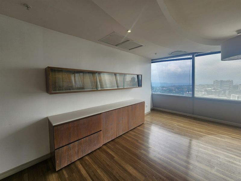 A bright, empty office space featuring hardwood flooring, built-in wooden cabinetry, and expansive windows offering panoramic city views.