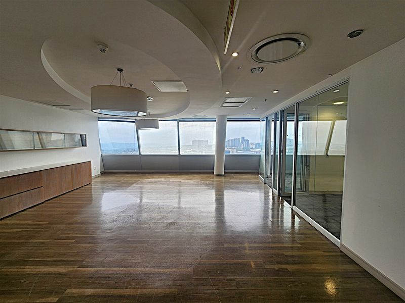 Spacious modern office area featuring hardwood flooring, floor-to-ceiling windows with city views, glass partitions, and integrated ceiling lighting.