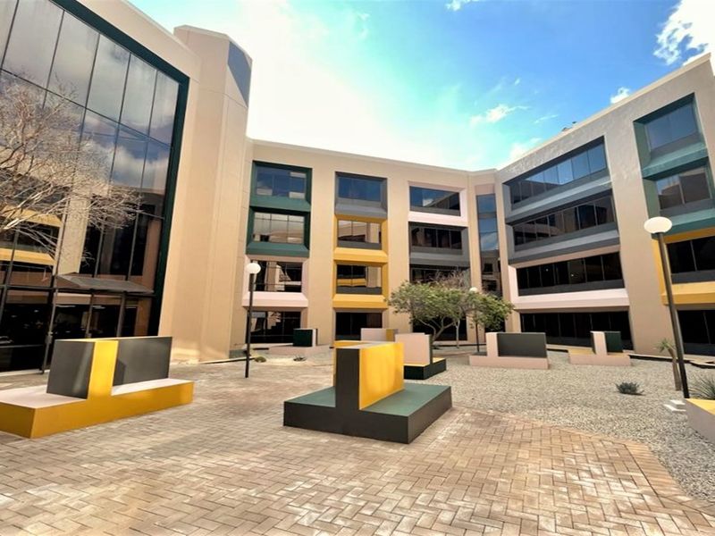 A modern office building courtyard featuring paved walkways, fixed concrete seating, and extensive glass curtain walling, providing a professional outdoor break area.