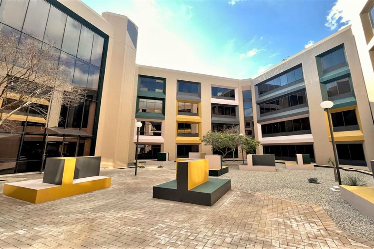 A modern office building courtyard featuring paved walkways, fixed concrete seating, and extensive glass curtain walling, providing a professional outdoor break area.