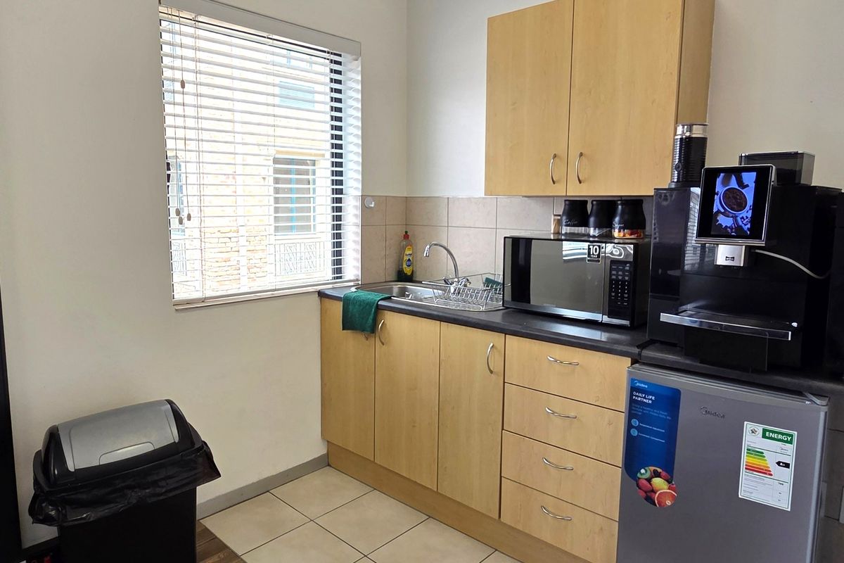 A compact office kitchenette featuring light wood-finish cabinetry, a stainless steel sink, tiled splashback, and tiled flooring.