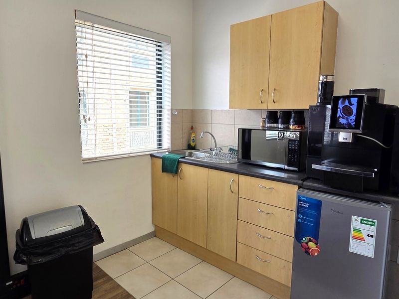 A compact office kitchenette featuring light wood-finish cabinetry, a stainless steel sink, tiled splashback, and tiled flooring.