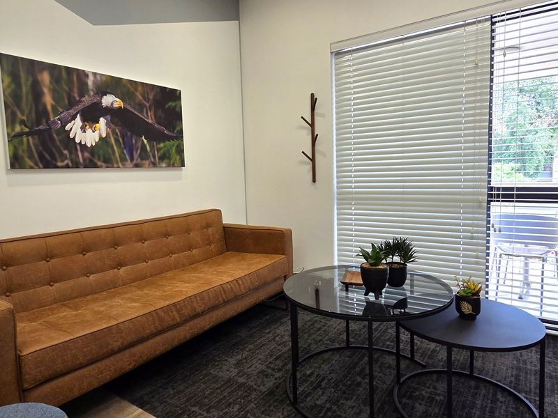 A professional office space featuring neutral walls, large windows with venetian blinds, and a wall-mounted coat rack.