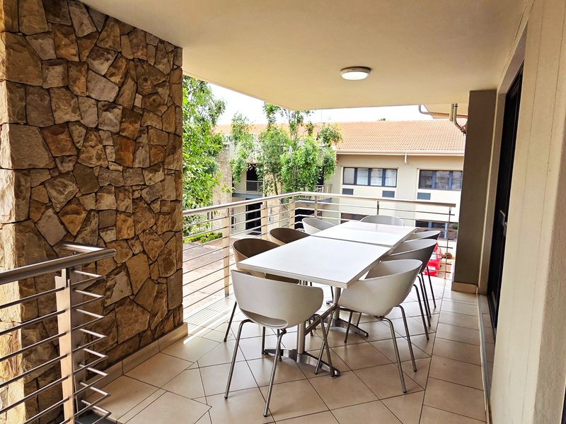 A covered balcony featuring a stone-clad feature wall, stainless steel railings, and tiled flooring, overlooking the surrounding office complex.