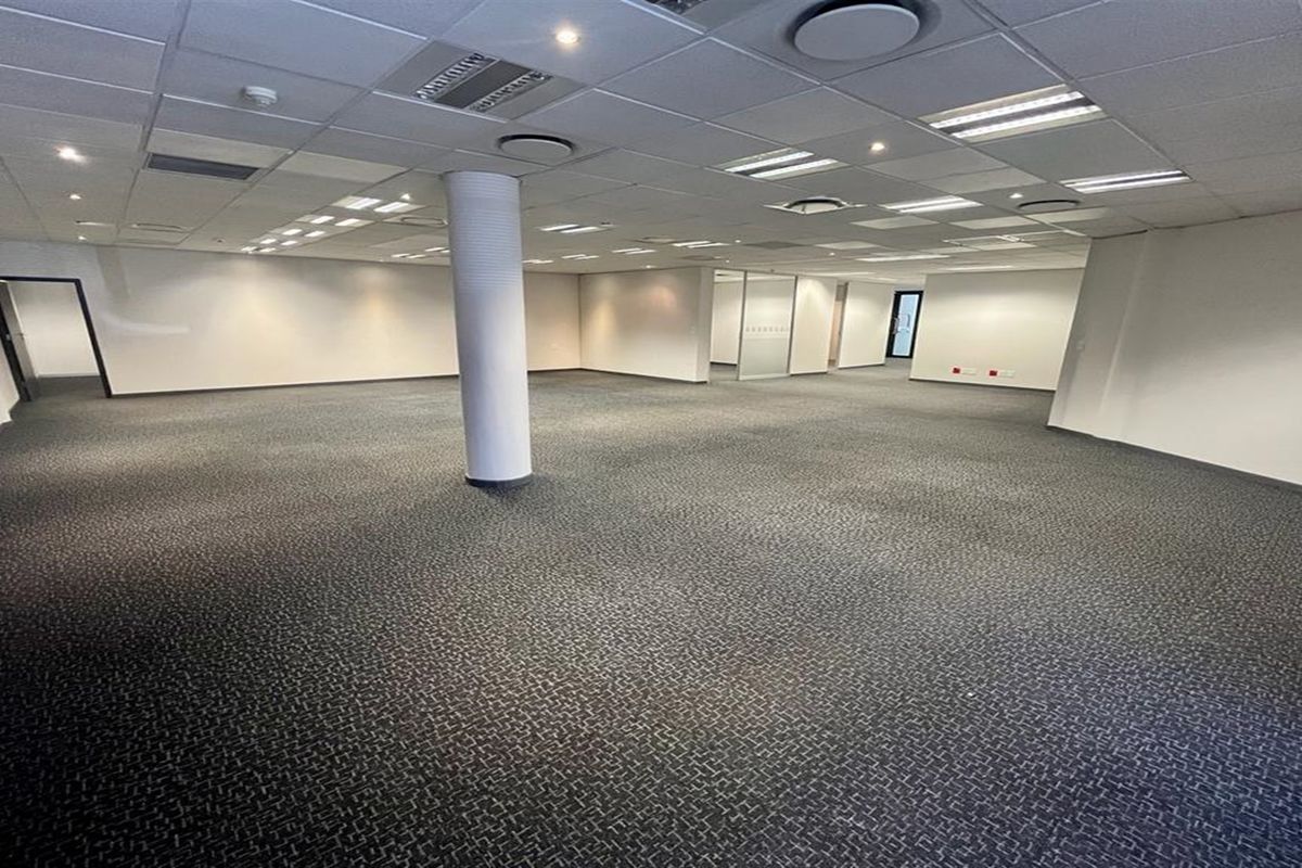 A spacious, open-plan office area featuring a suspended ceiling with recessed lighting, carpeted flooring, and a central structural column.