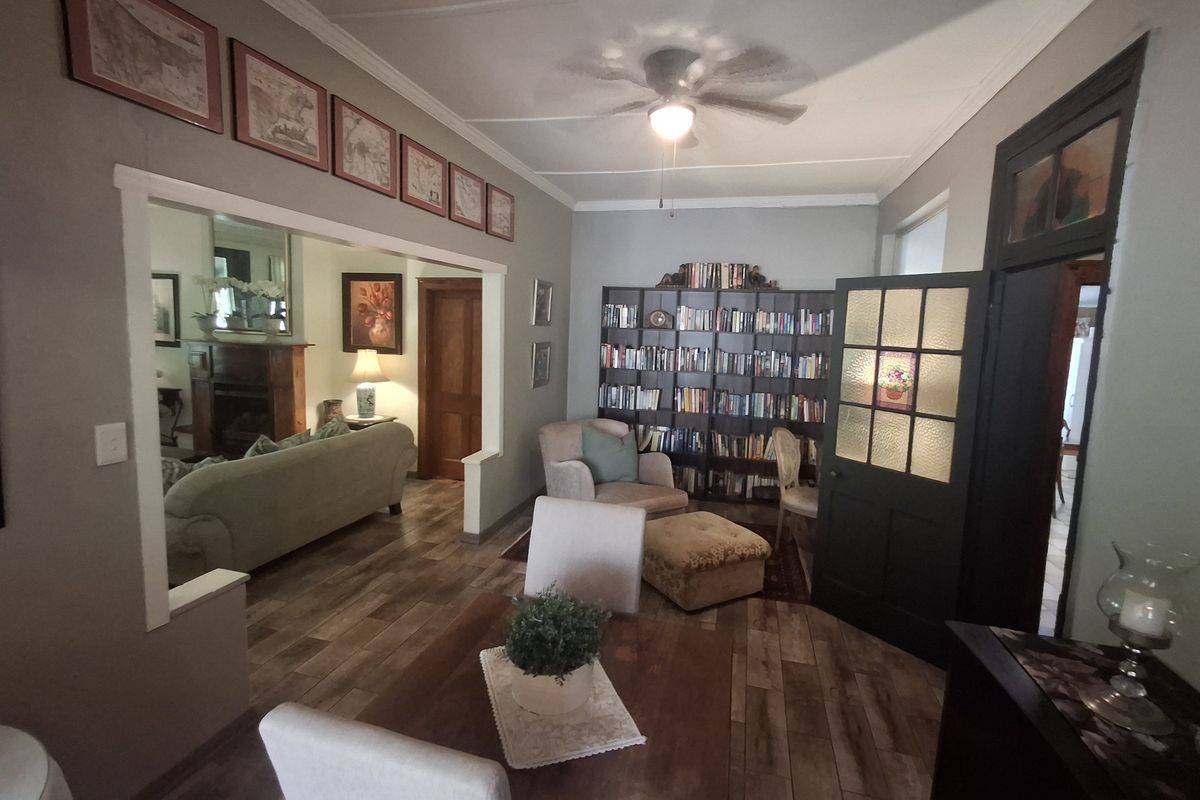 A cozy living area featuring timber-look flooring, a ceiling fan, and built-in shelving, with an open-plan flow into an adjacent room.