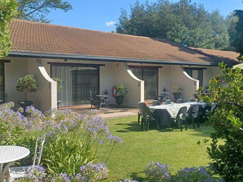 Rear view of the guest house featuring a well-maintained garden, paved patio areas with sliding door access to individual units, and a tiled roof.