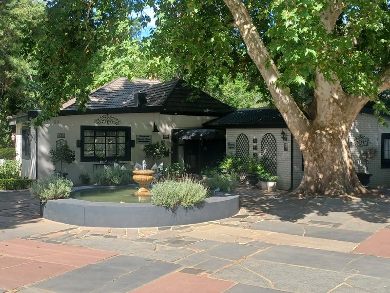 Front exterior view of the guest house featuring a stone paved driveway, central water fountain, and mature trees providing shade to the building facade.