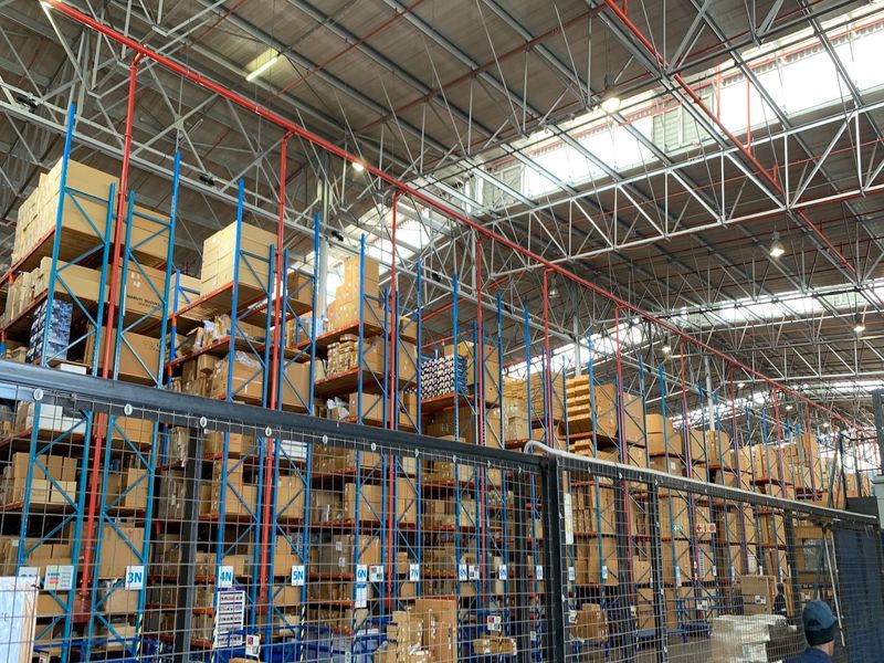 Large industrial warehouse space featuring extensive steel racking systems, exposed roof trusses, integrated fire sprinkler infrastructure, and natural light via overhead skylights.