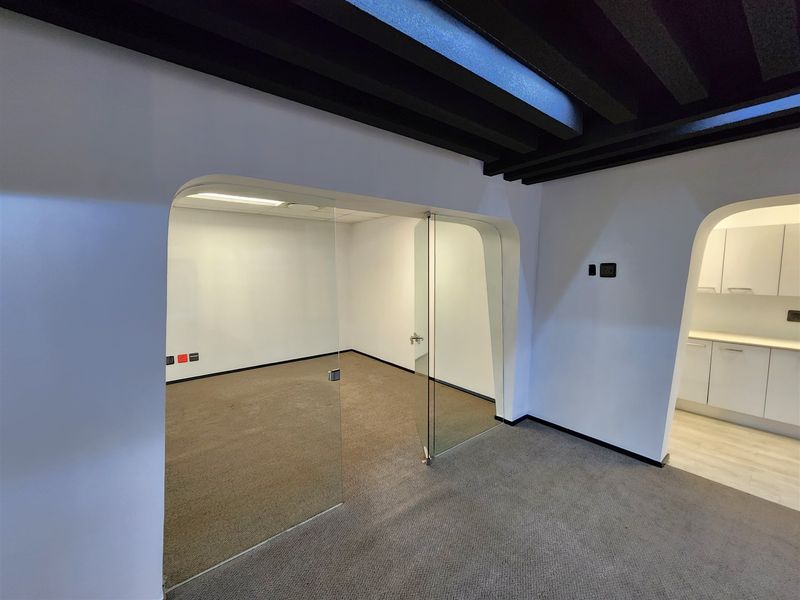 A modern, empty office space featuring glass pivot doors, arched architectural details, exposed ceiling beams, and an adjacent kitchenette area.
