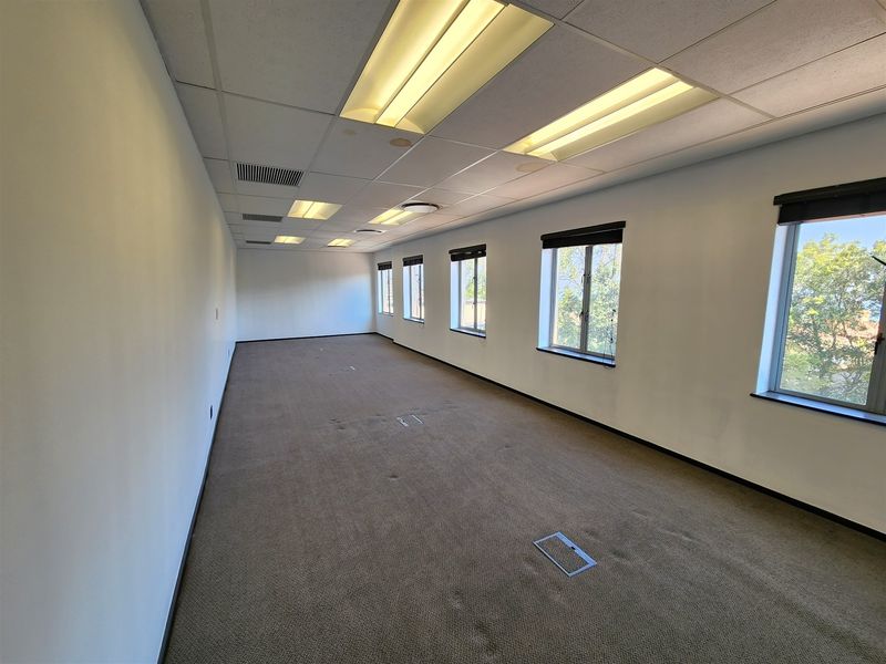 A spacious, empty office area featuring carpeted flooring, recessed lighting, and multiple windows providing natural light.