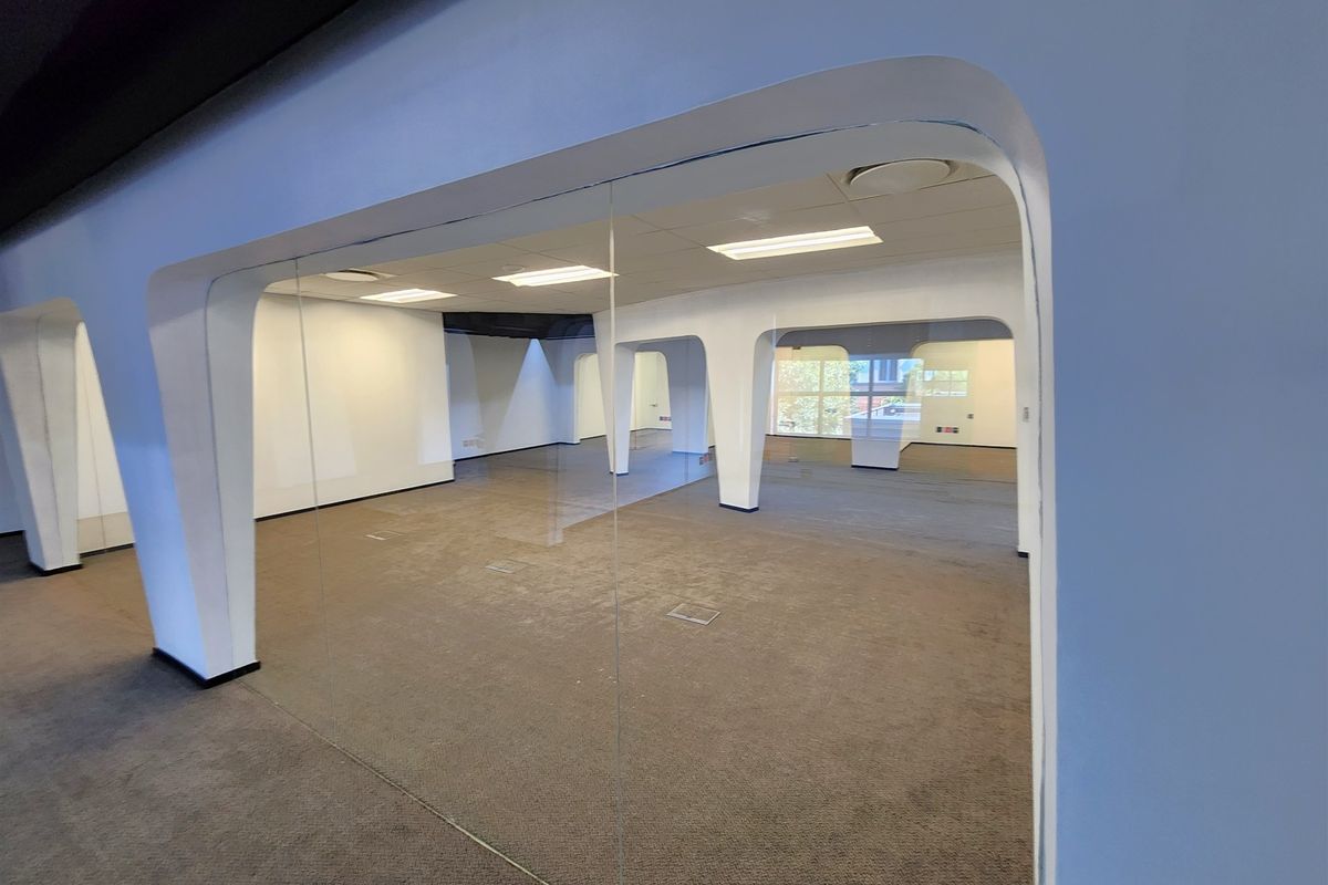 A spacious, open-plan office area featuring glass partitions, suspended ceilings with recessed lighting, and carpeted flooring with integrated floor power outlets.