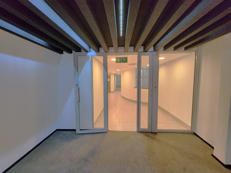 A clean, modern office hallway featuring glass partition doors, tiled flooring, and an exposed beam ceiling with integrated recessed lighting.