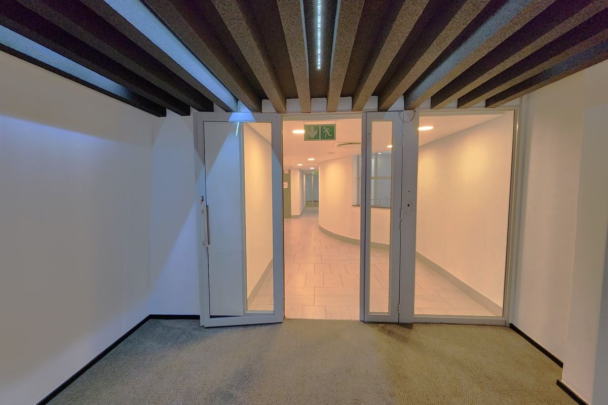 A clean, modern office hallway featuring glass partition doors, tiled flooring, and an exposed beam ceiling with integrated recessed lighting.