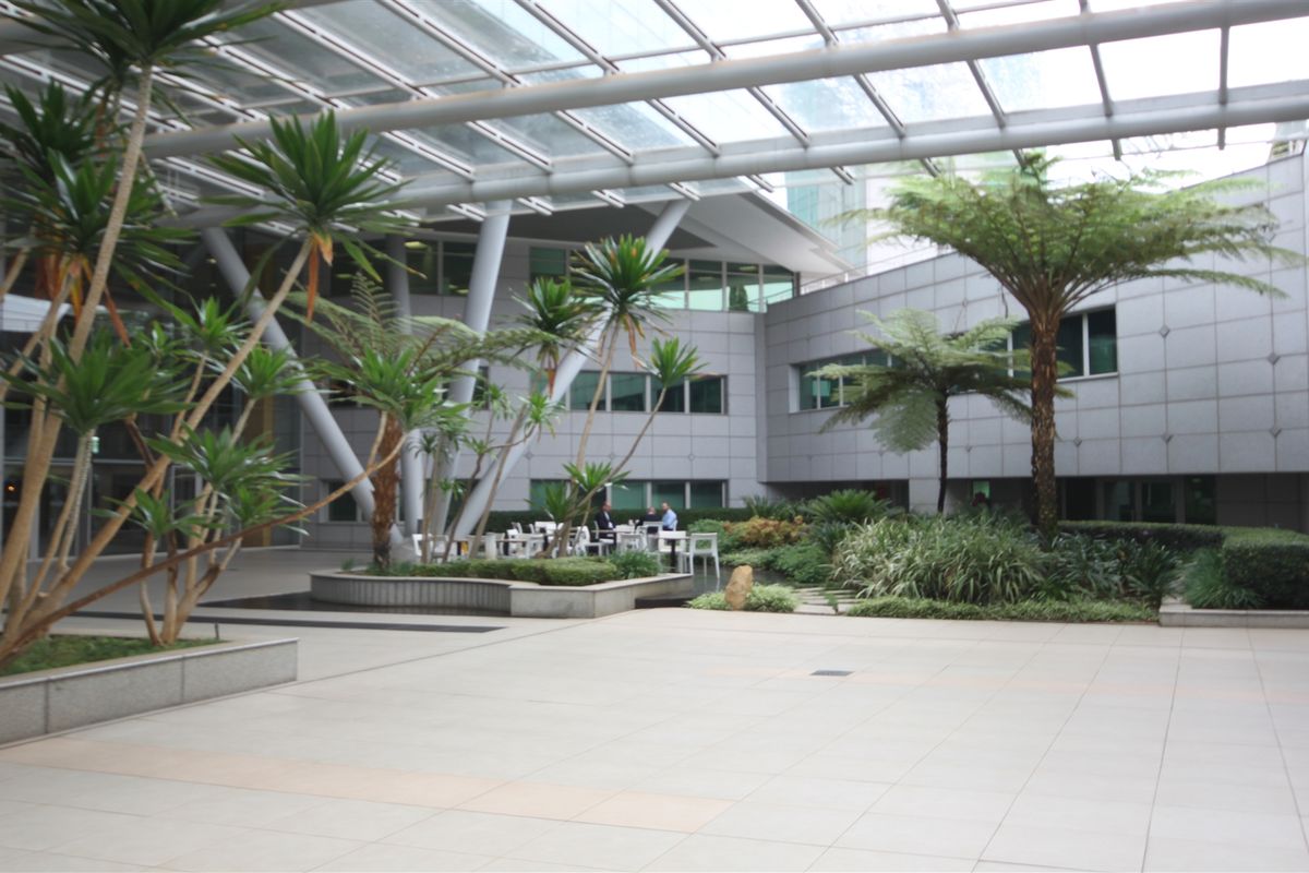A spacious, modern office courtyard featuring a glass atrium roof, tiled flooring, and integrated landscaped planter beds.