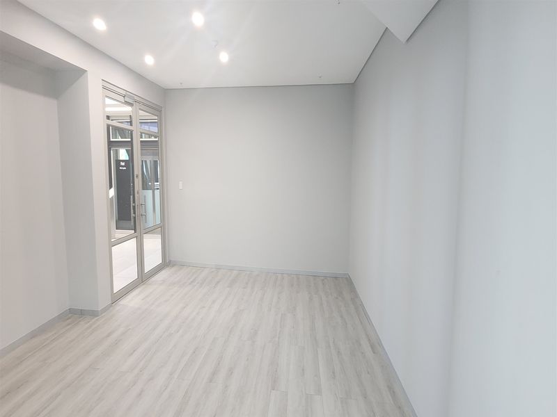 A bright, modern, and empty office space featuring light-toned laminate flooring, recessed ceiling lighting, and a glass-paneled entrance door.