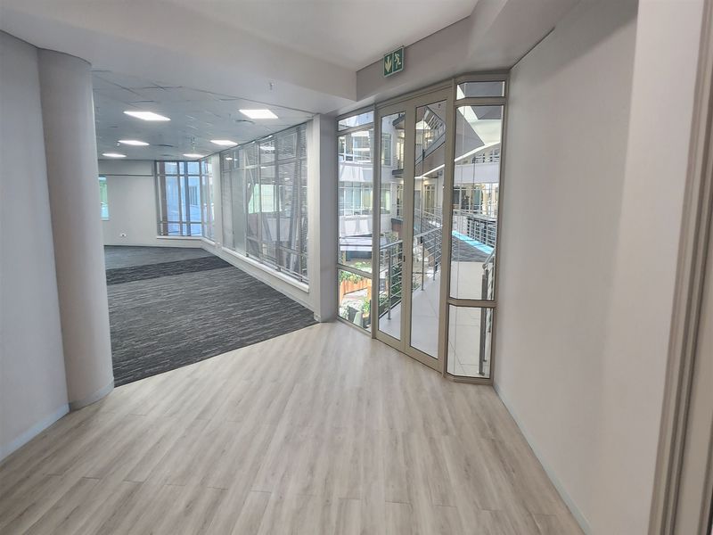 A bright, modern open-plan office space featuring a mix of laminate and carpet flooring, large windows, and direct access to an internal courtyard.