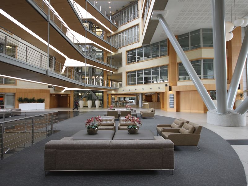 Spacious modern office atrium featuring a multi-level mezzanine, striking structural steel columns, and extensive glass facades providing abundant natural light.