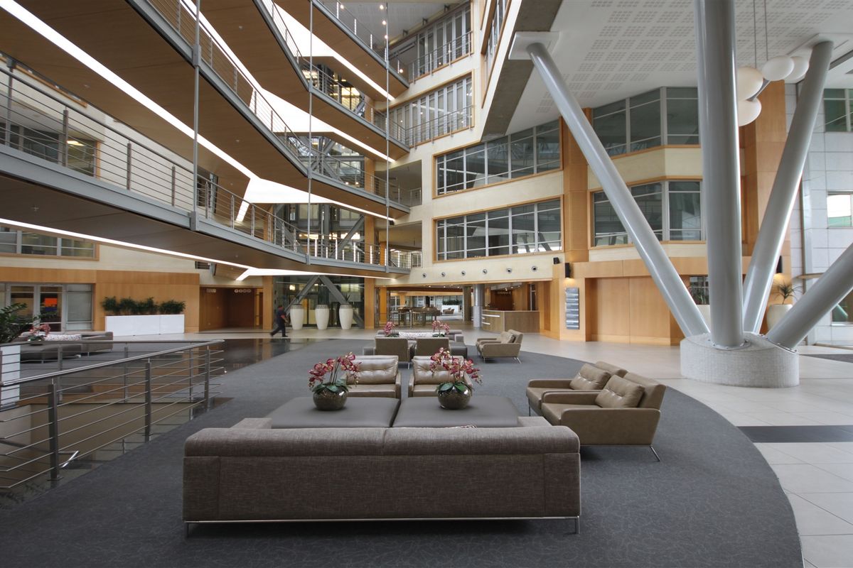Spacious modern office atrium featuring a multi-level mezzanine, striking structural steel columns, and extensive glass facades providing abundant natural light.