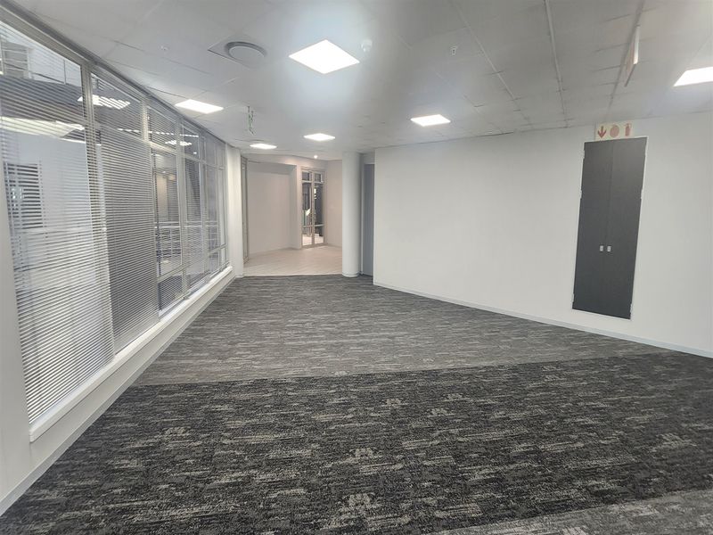 A spacious, empty open-plan office area featuring carpeted flooring, a suspended ceiling with recessed lighting, and large glass partitions.