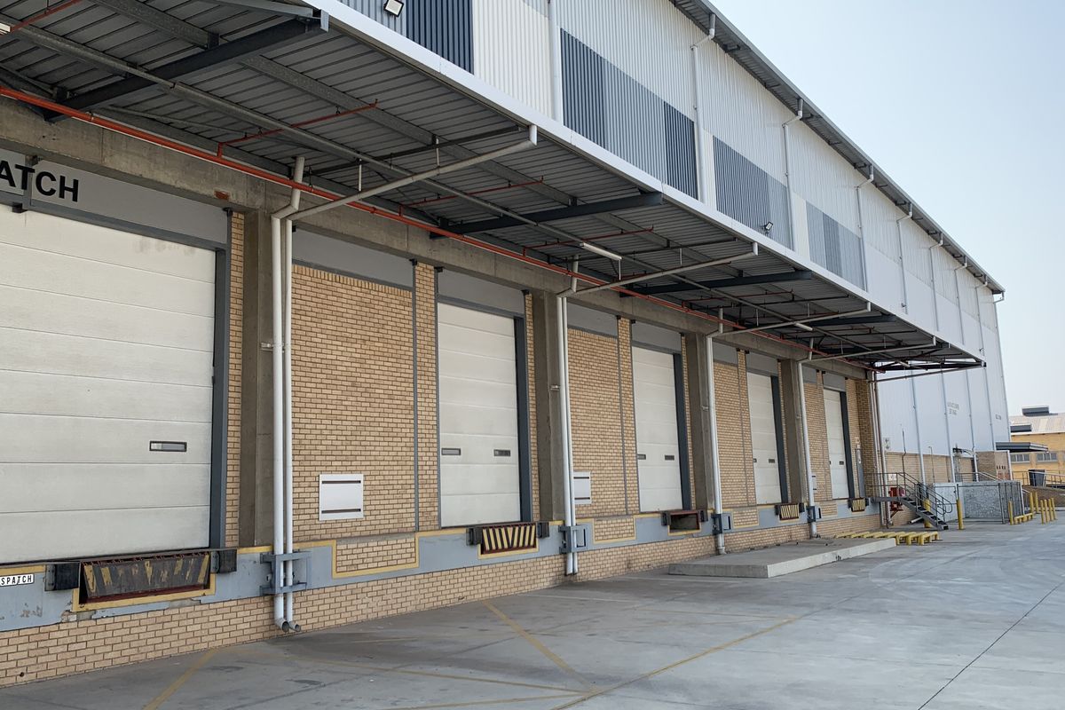 Industrial warehouse exterior featuring multiple loading docks with roller shutter doors, a protective metal canopy, and a spacious concrete yard area.