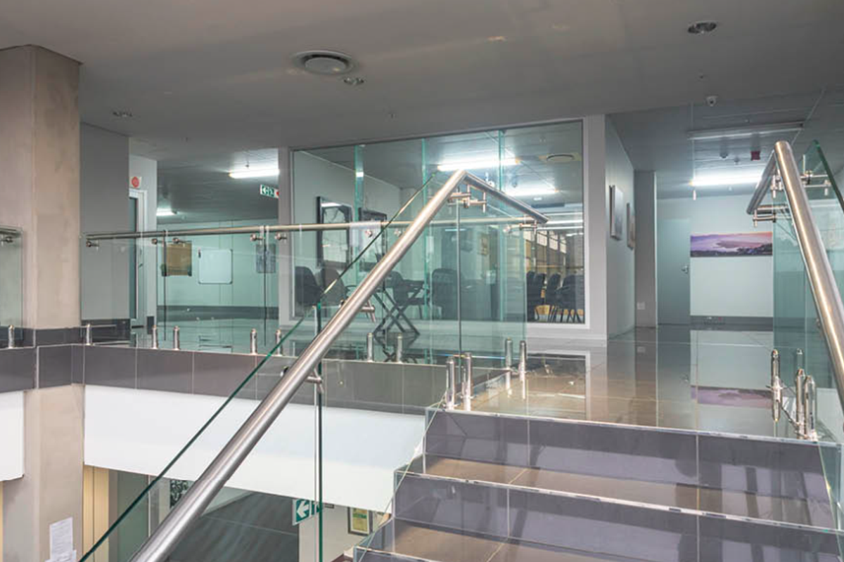 Modern industrial office hallway featuring a glass balustrade staircase, tiled flooring, and glass-partitioned office spaces with recessed lighting.